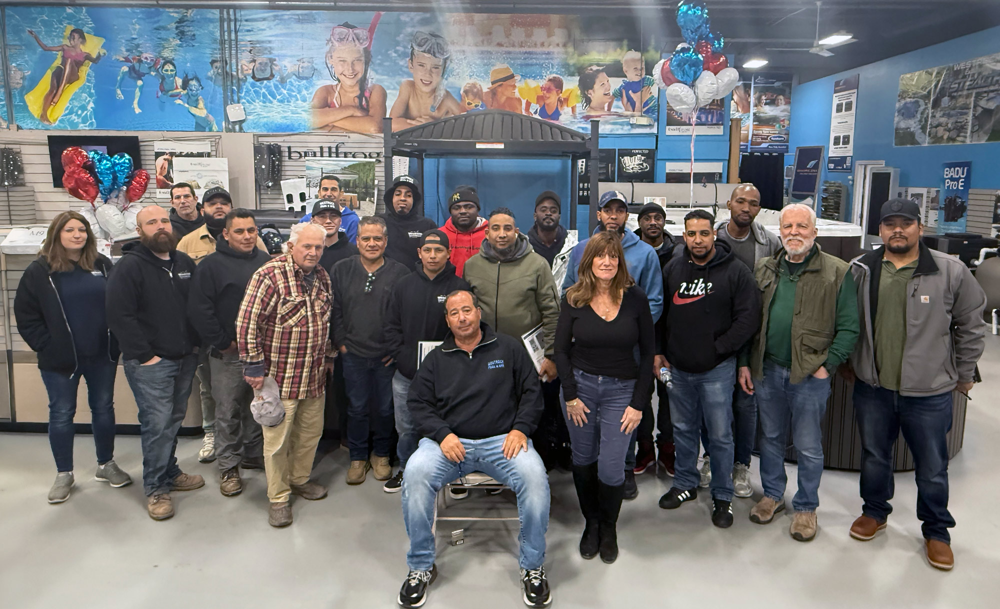 Group of about 20 adults posing for a team photo inside a store showroom with a blue mural and balloons in the background.