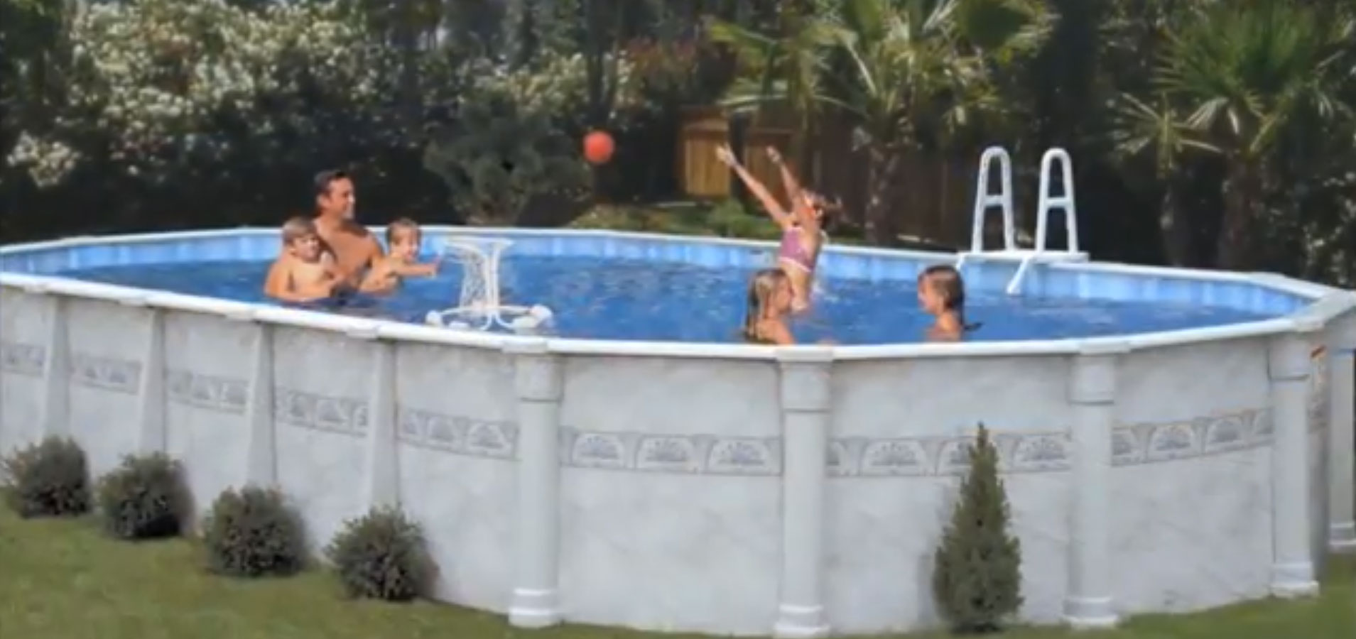 Group of kids and an adult in an above-ground pool; a girl in pink jumps with arms raised to catch an orange ball.