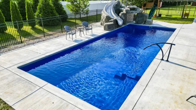 Rectangular in-ground pool on a concrete deck with a stone water slide at one end, a metal ladder, and two chairs nearby; green lawn and trees in the background.