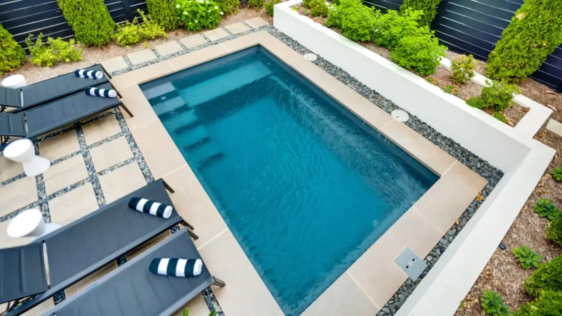 Rectangular backyard pool with turquoise water, beige pool deck, and several black lounge chairs with striped towels along the side.