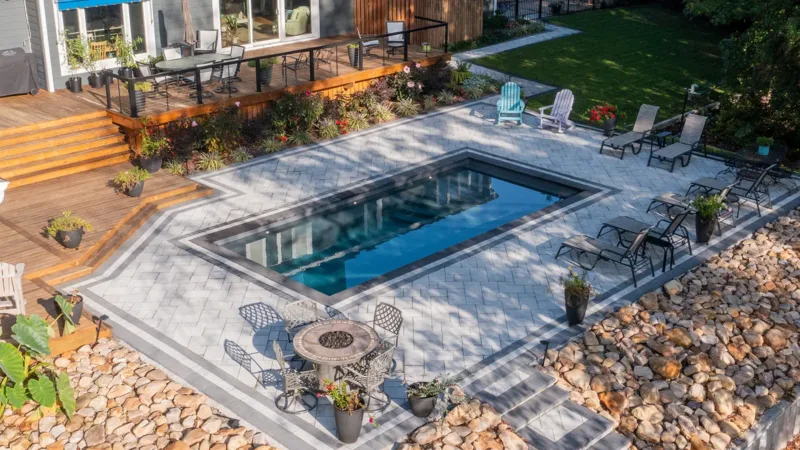 Rectangular in-ground pool with a gray stone patio, surrounding lounge chairs, a wooden deck with seating, and landscaped rocks in a modern backyard.