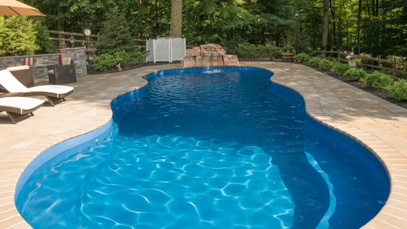 Outdoor kidney-shaped pool with bright blue water, beige stone deck, two lounge chairs under an umbrella, and a wooded backyard with trees in the background.