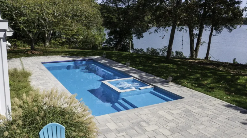 Rectangular pool with an attached square spa, set in a light stone patio, with trees and a lake in the background.