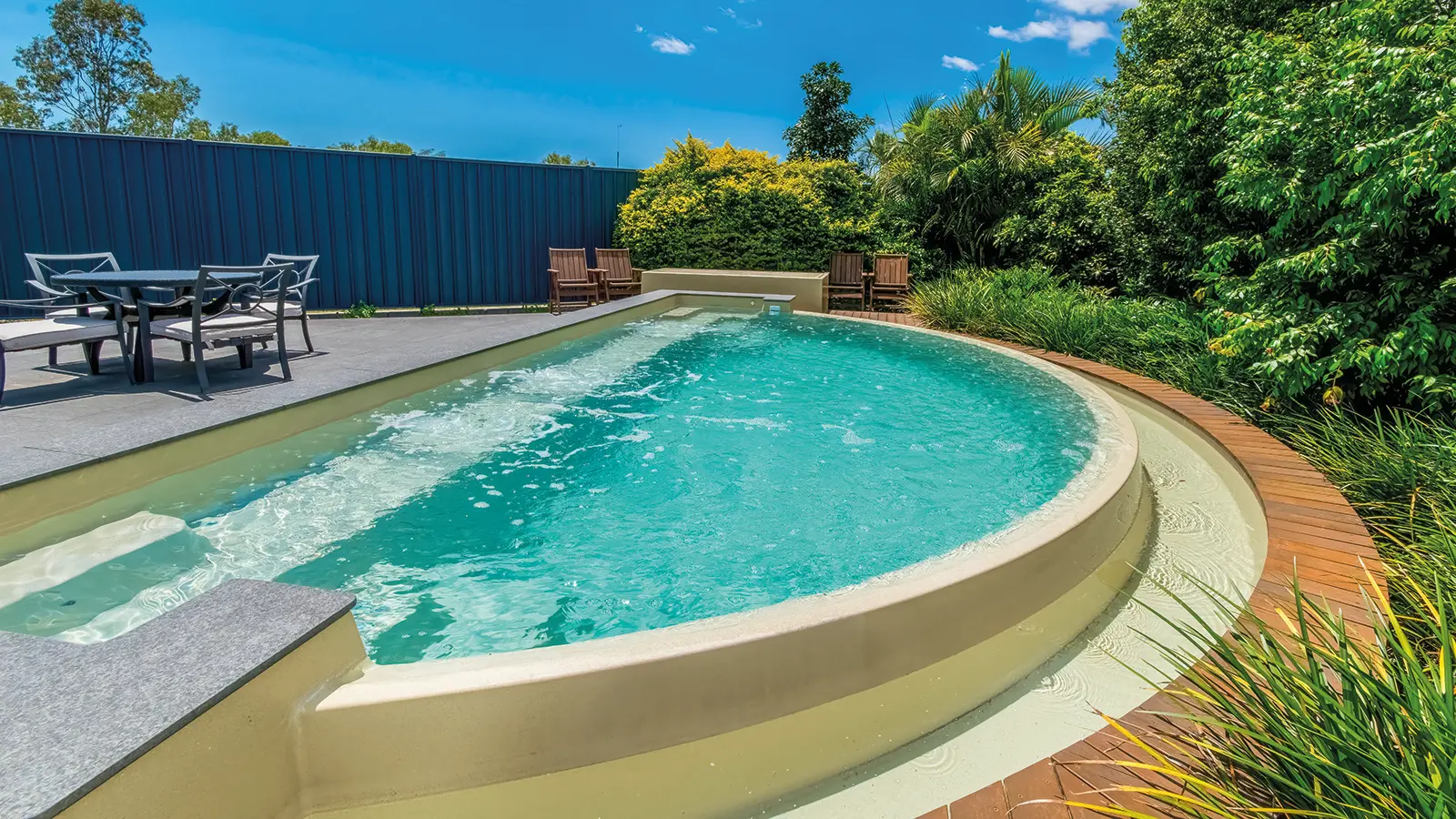 Curved turquoise pool in a sunny backyard, with a wood deck, outdoor seating, and dense green shrubs along a blue fence.