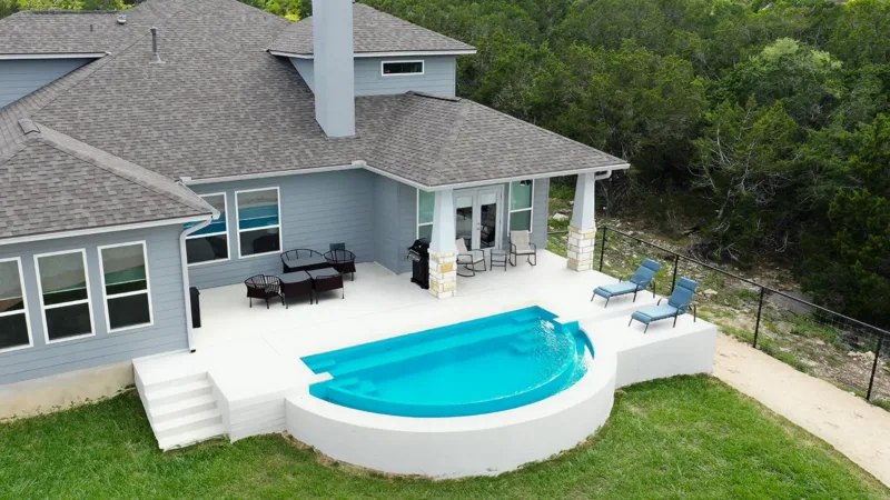 Modern gray house with a curved in-ground blue pool on a white deck, seating area, and a covered patio; trees in the background.