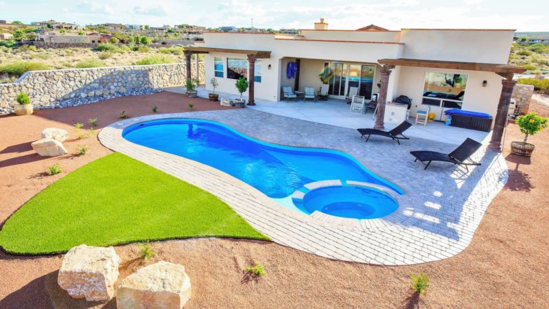 Modern single-story home with a curved blue swimming pool and round hot tub, patio lounge chairs, and desert landscaping overlooking a rocky landscape.