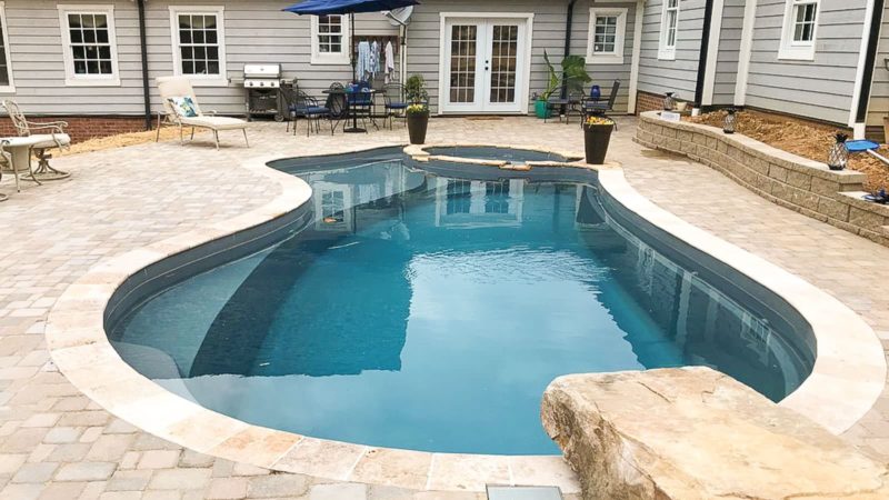 Curved in-ground swimming pool in a paved backyard with outdoor seating, a grill, and a house with French doors in the background.