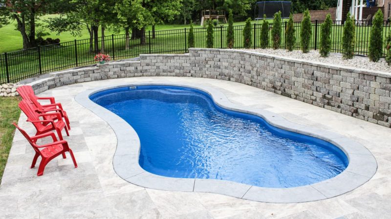 Curved blue pool in a backyard with a gray stone patio and red chairs along the edge.