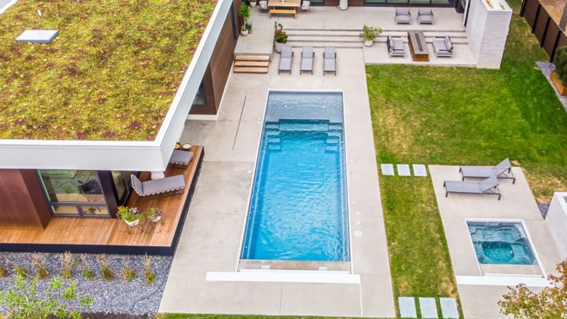 Aerial view of a modern backyard with a long rectangular pool, hot tub, and lounge area beside a green lawn and wood deck.