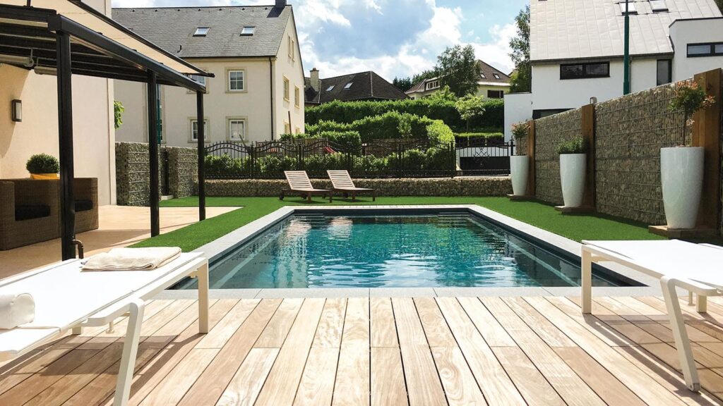 Modern backyard with rectangular pool, wooden deck, and two white lounge chairs on a green lawn; stone walls and large planters line the fence before neighboring houses.