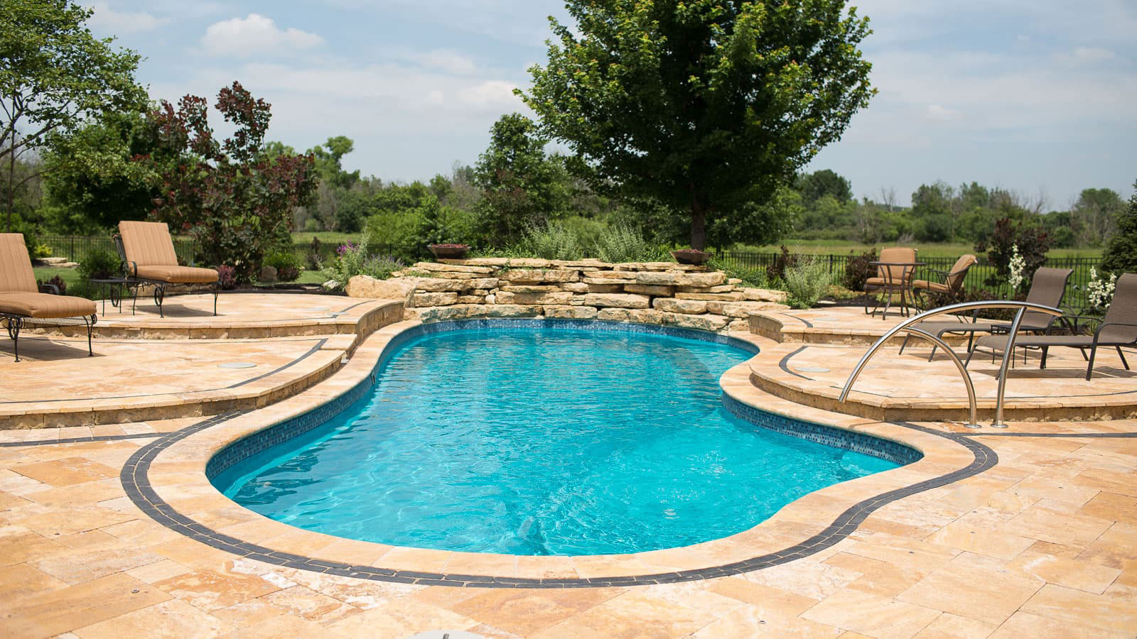Curved in-ground pool surrounded by tan stone patio and lounge chairs in a sunny backyard.
