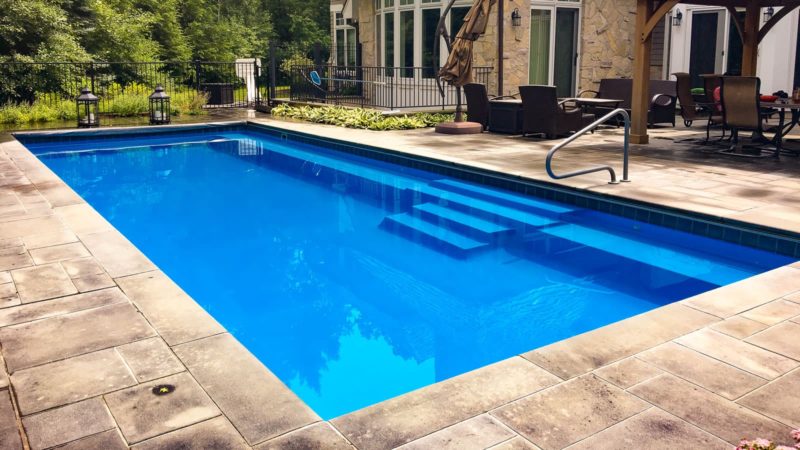 Blue rectangular backyard pool with built-in steps and a metal handrail, surrounded by a stone patio and outdoor seating.