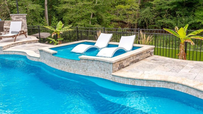 Outdoor pool with a raised spa and two white lounge chairs on a stone deck, surrounded by greenery and railing