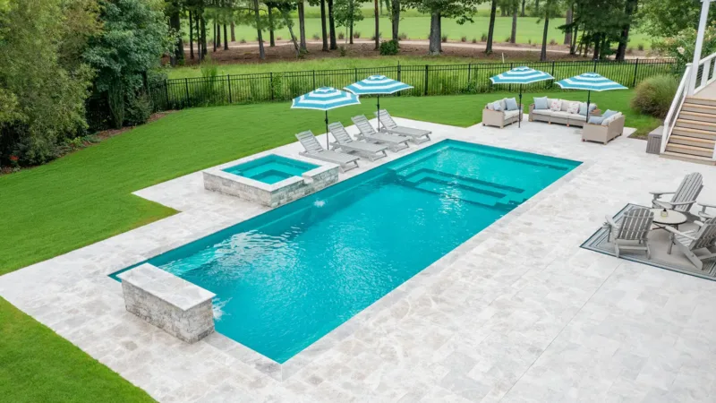 Rectangular in-ground pool with a raised spa, surrounded by a light stone deck and green lawn on a sunny backyard.