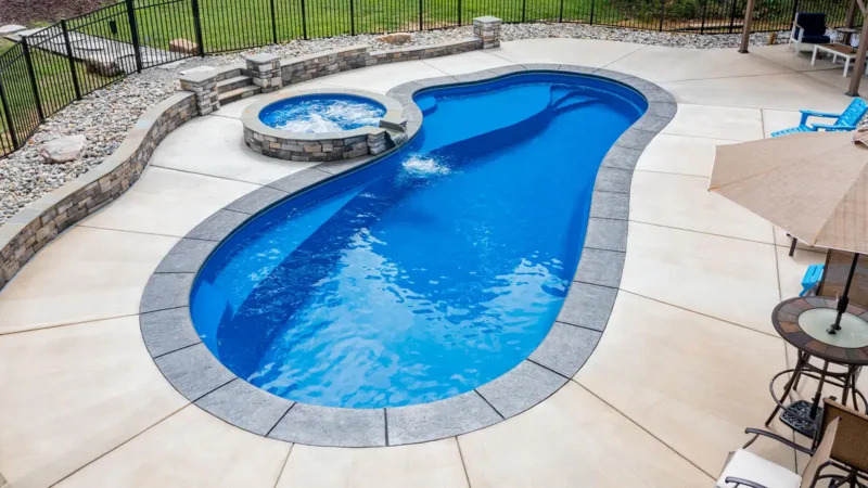 Curved blue in-ground pool with attached circular hot tub, surrounded by gray stone coping and a beige concrete deck.