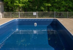 Blue empty swimming pool with white tiled rim, fence with a 'No Diving' sign, and dense green trees in the background.