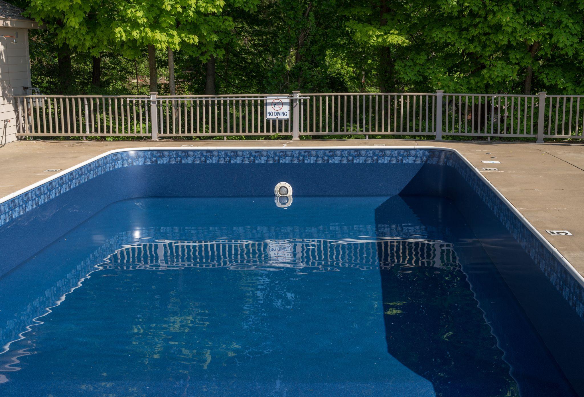 Blue empty swimming pool with white tiled rim, fence with a 'No Diving' sign, and dense green trees in the background.