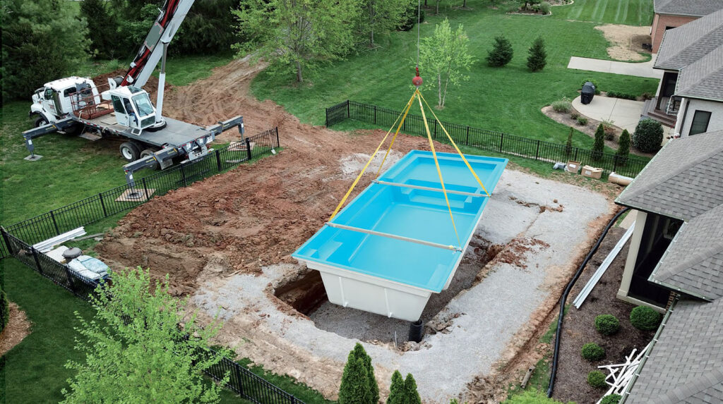Backyard construction scene: a crane lowers a blue in-ground pool module into a wide trench, with dirt, gravel, and a concrete pad; a truck-mounted crane and fencing surround the yard near a neighboring house.
