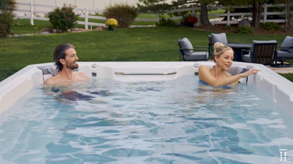 Two people relaxing in an outdoor white hot tub with a grassy yard and patio seating in the background.
