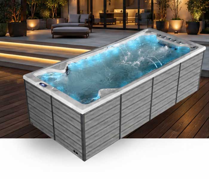 Rectangular outdoor hot tub on a wooden deck with blue water and a nighttime lounge area in the background.