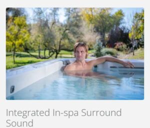 Man relaxing in an outdoor hot tub, with water and greenery in a sunny garden setting, showcasing in-spa surround sound feature