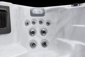 White hot tub interior featuring an array of chrome jet nozzles arranged in two vertical columns, with a small vent on the left and a rounded control panel above.