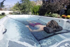 Person with long hair lounging on a black pool pillow while floating in a clear blue backyard pool, trees and mountains in the background.