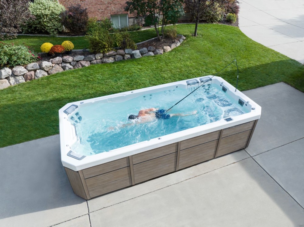 Person swimming in a rectangular outdoor hot tub on a concrete patio beside a green lawn and stone-edged garden bed.