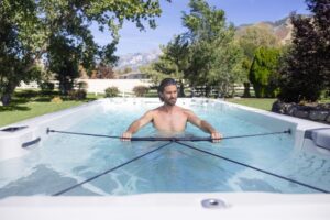 Shirtless man rowing in a rectangular backyard pool with two long oars attached to the pool rails, mountains and trees in the background