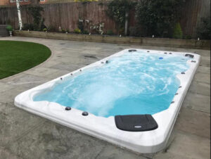 White rectangular outdoor hot tub with bubbling blue water on a stone patio, backyard with a wooden fence in the background