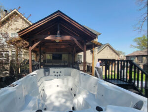 White hot tub in a suburban backyard under a dark wooden pavilion with a deck; a man stands nearby on the deck.