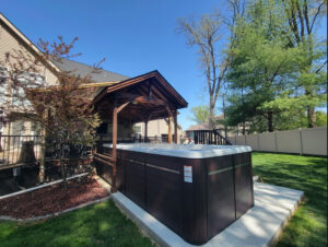 Backyard with a large black hot tub under a wooden pavilion attached to the house, with green grass and trees nearby in bright daylight.