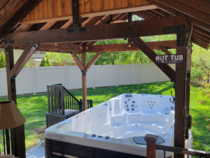 Wooden gazebo-style structure over a white hot tub in a green backyard with a white fence behind it.