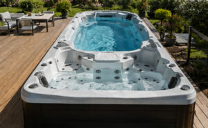 Large white outdoor hot tub with built-in seating and jets on a wooden deck, with a garden and patio furniture nearby.
